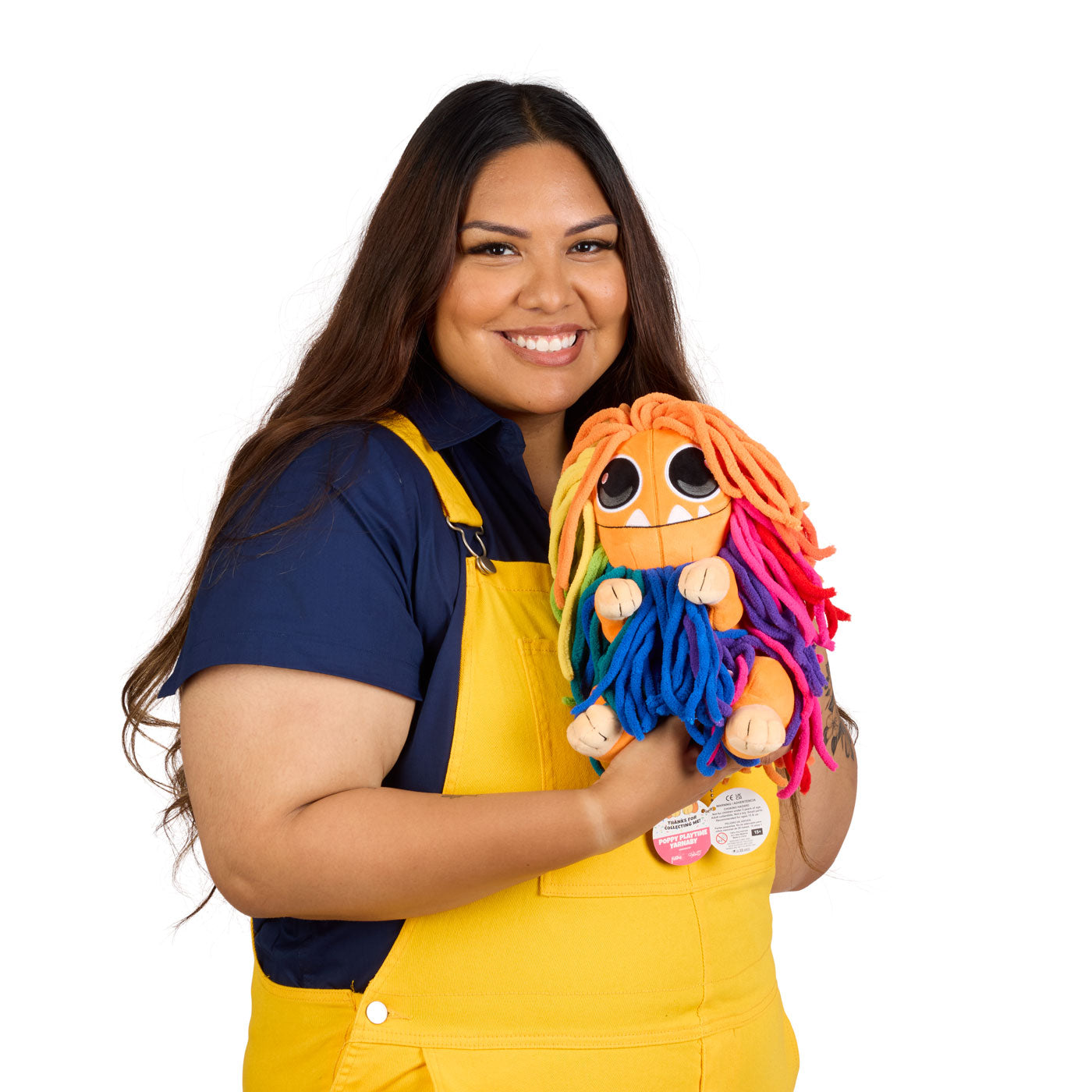 Model wearing a navy blue shirt and yellow overalls, holds a colorful plush toy with bright orange, pink, blue, and purple yarn-like hair.