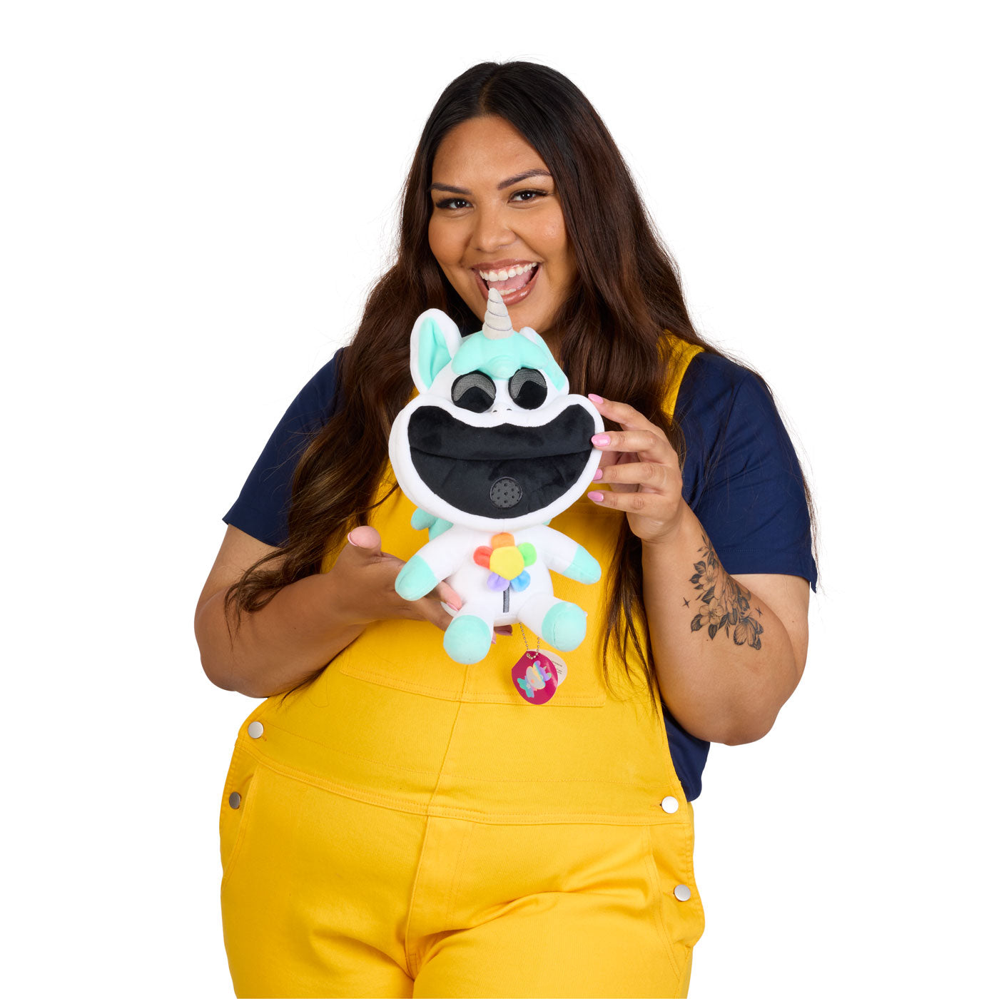 A model holding a plush unicorn toy. She is wearing yellow overalls over a navy blue t-shirt.